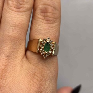 以下が含まれることがあります： 中央にエメラルドカットの緑色の宝石をあしらったゴールドリング。周囲には小さなクリアストーンが配置されています。リングは幅広のバンドと装飾的なセッティングが特徴で、クラシックなジュエリーデザインを示しています。