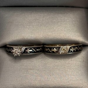 May include: Two silver rings with black enamel detailing and cross designs. One ring features a central diamond, the other has small accent stones. The rings are displayed in a grey ring box.