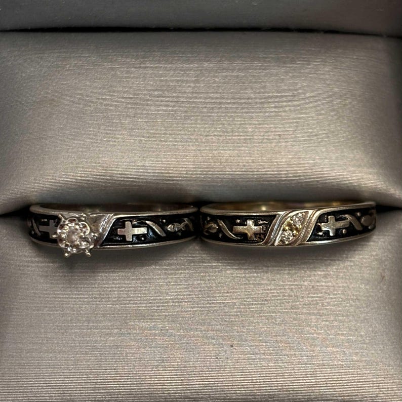 May include: Two silver rings with black enamel detailing and cross designs. One ring features a central diamond, the other has small accent stones. The rings are displayed in a grey ring box.
