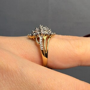 May include: A gold ring featuring a cluster of small, sparkling diamonds. The ring has a split shank design and is set against a neutral background. The diamonds are clustered together, creating a floral or starburst effect.