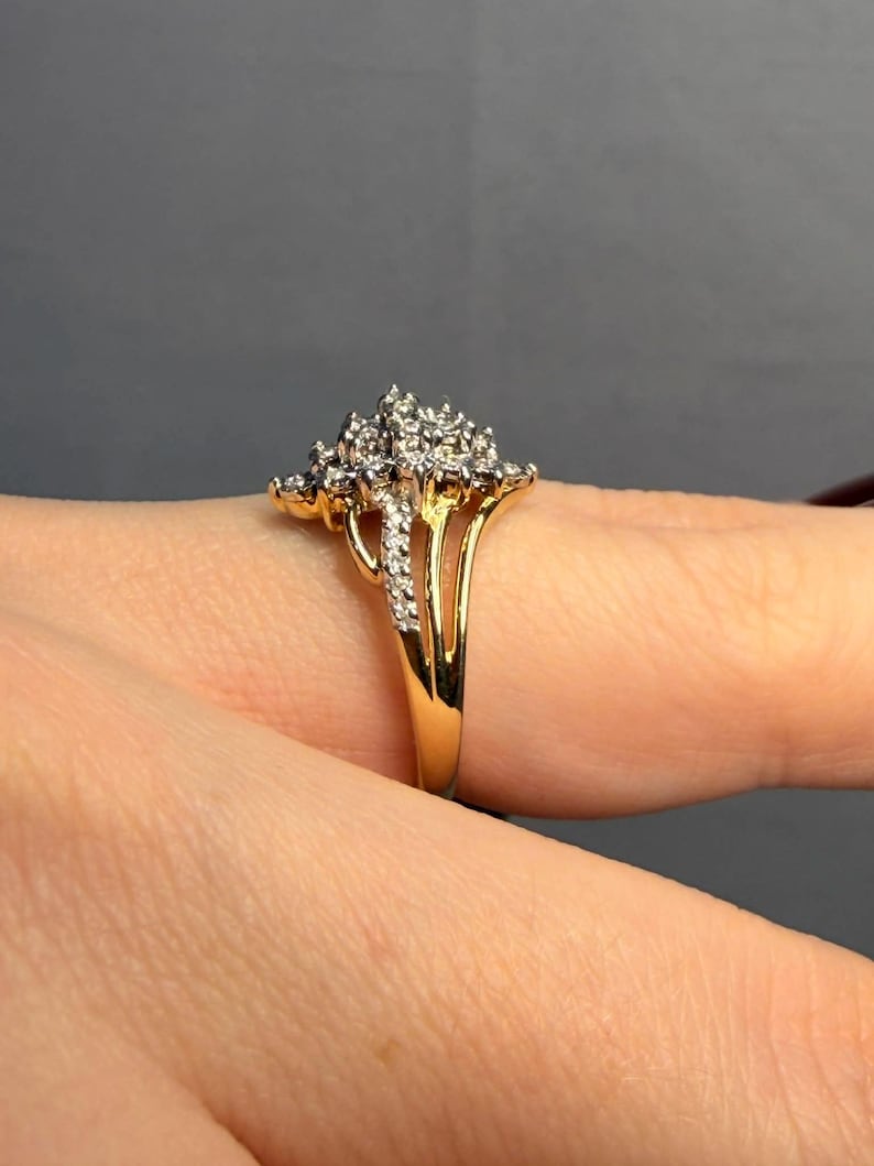 May include: A gold ring featuring a cluster of small, sparkling diamonds. The ring has a split shank design and is set against a neutral background. The diamonds are clustered together, creating a floral or starburst effect.
