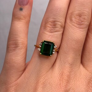 May include: A gold ring featuring a large, rectangular emerald-cut green gemstone. Small, clear accent stones surround the main stone. The ring is set on a gold band, showcasing a classic and elegant design.