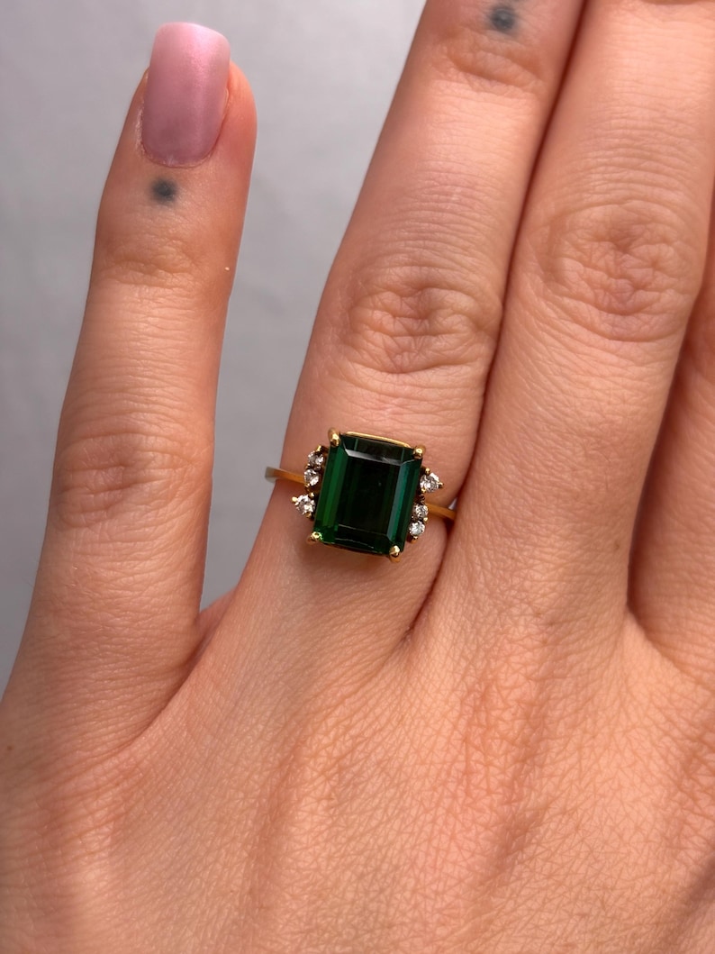 May include: A gold ring featuring a large, rectangular emerald-cut green gemstone. Small, clear accent stones surround the main stone. The ring is set on a gold band, showcasing a classic and elegant design.