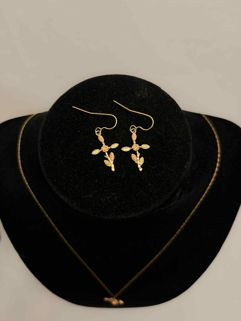 Pode incluir: Conjunto de joias douradas com um par de brincos em forma de cruz e um colar. Os brincos e o pingente t&ecirc;m um design floral. As joias s&atilde;o exibidas em uma superf&iacute;cie de veludo preto.
