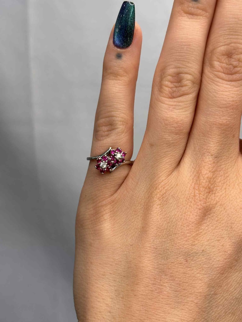 May include: A silver ring featuring a cluster of small, round, red gemstones and a single white gemstone. The gemstones are set in a floral-like arrangement. The ring is on a finger with a dark teal nail polish.