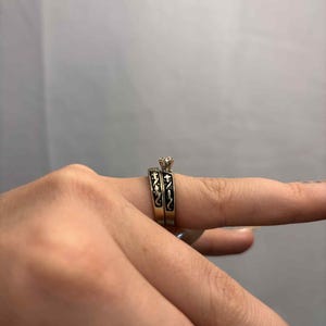 May include: A two-piece wedding ring set. The engagement ring features a small, clear gemstone. The bands are gold-toned with black enamel detailing. The rings are on a finger.
