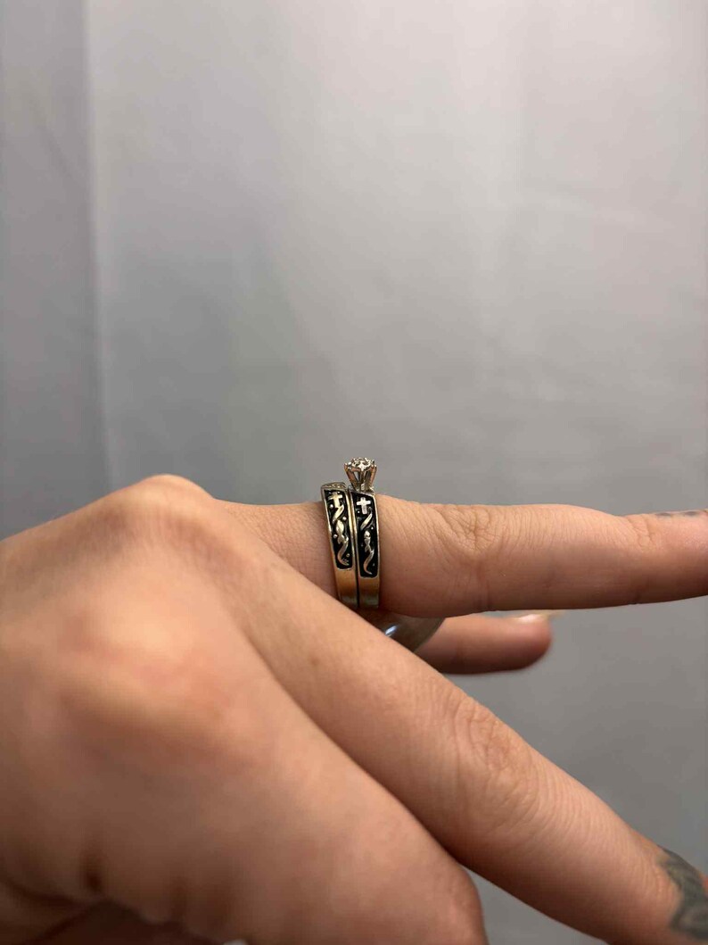 May include: A two-piece wedding ring set. The engagement ring features a small, clear gemstone. The bands are gold-toned with black enamel detailing. The rings are on a finger.