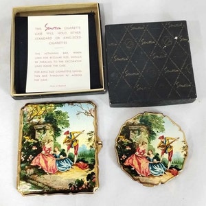 May include: A vintage Stratton cigarette case with a gold trim and a painted scene of a couple in a garden. The case is closed and the lid has a hinge. The case is in its original box, which is black with a Stratton logo.