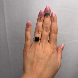May include: A gold ring featuring a large, square, dark green gemstone. The ring has a delicate, ornate setting. The hand wearing the ring has pink nail polish and small tattoos.
