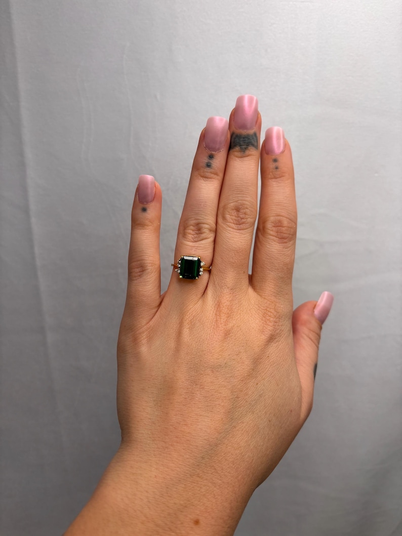 May include: A gold ring featuring a large, square, dark green gemstone. The ring has a delicate, ornate setting. The hand wearing the ring has pink nail polish and small tattoos.
