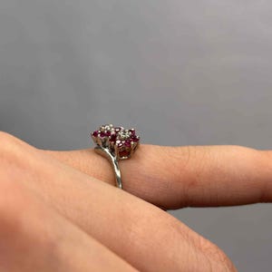 May include: A silver ring featuring a cluster of small, round, red gemstones surrounded by smaller, clear stones. The ring is set against a neutral gray background, highlighting the jewelry's design and sparkle.