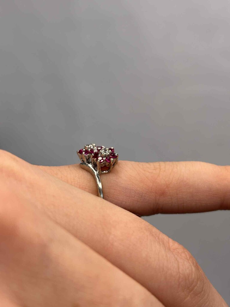 May include: A silver ring featuring a cluster of small, round, red gemstones surrounded by smaller, clear stones. The ring is set against a neutral gray background, highlighting the jewelry's design and sparkle.