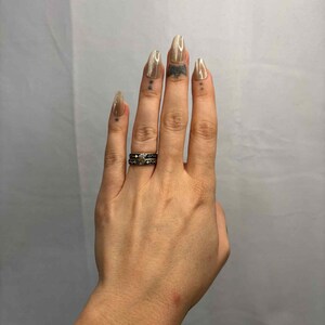 May include: Hand with long, almond-shaped, light brown nails. The hand has a silver ring with a dark design on the ring finger. There are small tattoos on the fingers.