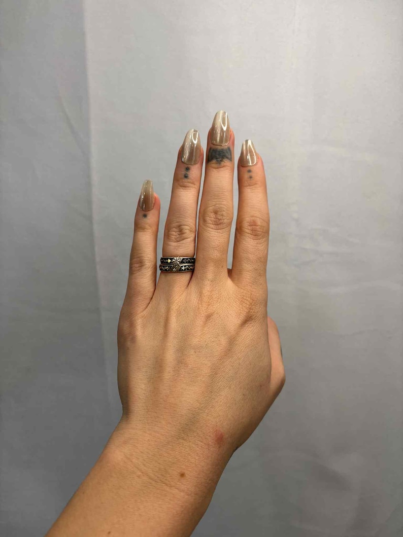 May include: Hand with long, almond-shaped, light brown nails. The hand has a silver ring with a dark design on the ring finger. There are small tattoos on the fingers.