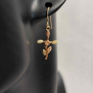 Pode incluir: Brinco dourado com um design de cruz floral. A cruz apresenta uma pequena flor no centro e detalhes de folhas. O brinco &eacute; suspenso por um gancho simples.