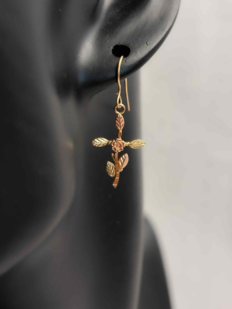 Pode incluir: Brinco dourado com um design de cruz floral. A cruz apresenta uma pequena flor no centro e detalhes de folhas. O brinco &eacute; suspenso por um gancho simples.