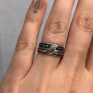 May include: Two rings are stacked on a finger. The top ring is gold with a diamond, and the bottom ring is black with gold accents. The rings have a cross design.