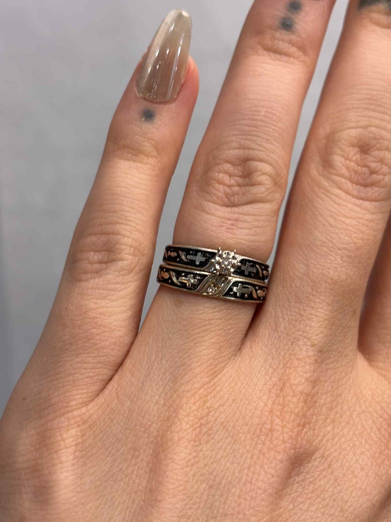 May include: Two rings are stacked on a finger. The top ring is gold with a diamond, and the bottom ring is black with gold accents. The rings have a cross design.