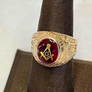 May include: A gold-colored ring with a large, oval, deep red stone. The ring features a Masonic symbol in gold and is engraved with text around the stone. The ring is displayed on a dark brown stand.