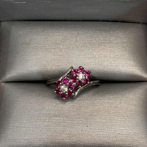 May include: A silver ring featuring two floral clusters. Each cluster has vibrant pink gemstones surrounding a central clear gemstone. The ring is set against a gray background, showcasing its elegant design.