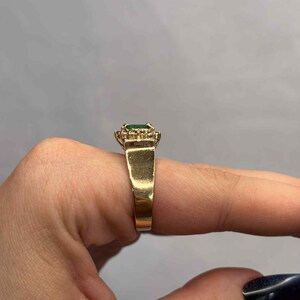 以下が含まれることがあります： 長方形のエメラルドカットの緑色の宝石をあしらったゴールドリング。周囲には小さなクリアストーンが配置されています。リングは太く磨かれたゴールドバンドです。指にはめられたリング。