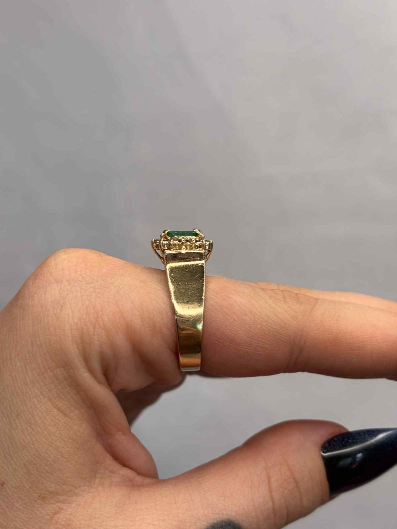 以下が含まれることがあります： 長方形のエメラルドカットの緑色の宝石をあしらったゴールドリング。周囲には小さなクリアストーンが配置されています。リングは太く磨かれたゴールドバンドです。指にはめられたリング。