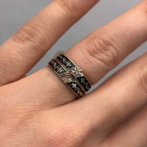 May include: A two-piece ring set with gold bands, black enamel detailing, and cross patterns. The engagement ring features a small, clear gemstone. The rings are displayed on a finger.
