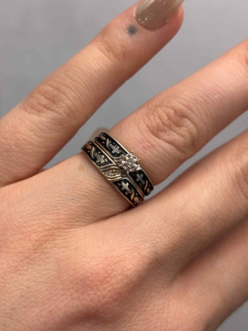 May include: A two-piece ring set with gold bands, black enamel detailing, and cross patterns. The engagement ring features a small, clear gemstone. The rings are displayed on a finger.