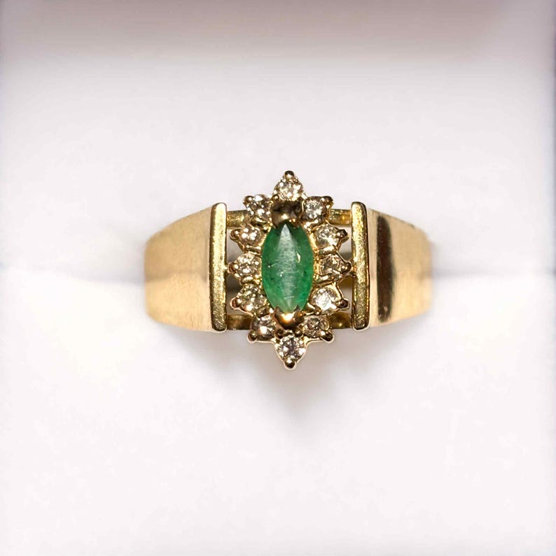 May include: A gold ring featuring a marquise-cut emerald center stone surrounded by a halo of small diamonds. The ring has a wide, flat band and a decorative setting. The emerald is a vibrant green, and the diamonds sparkle against the gold.