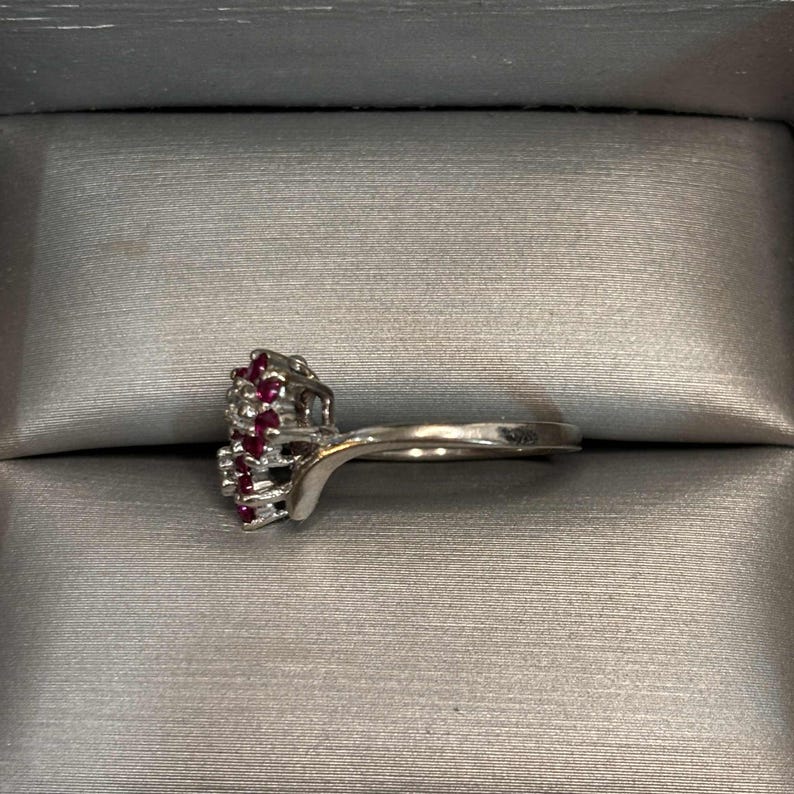 May include: A silver ring with a cluster of small, round, red gemstones. The ring is set against a gray background, showcasing the jewelry's design and color.