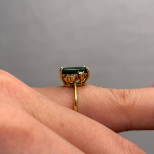 May include: A gold ring featuring a large, rectangular, dark green gemstone. The ring has a delicate gold band and is accented with small, clear gemstones. The ring is shown on a finger.
