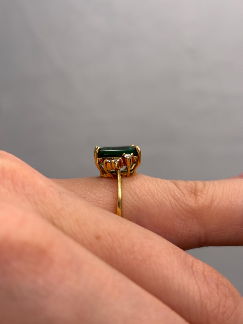 May include: A gold ring featuring a large, rectangular, dark green gemstone. The ring has a delicate gold band and is accented with small, clear gemstones. The ring is shown on a finger.