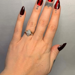 May include: A hand with long, oval-shaped, glossy red nails. A gold ring with a diamond cluster is on the ring finger. The nails have a deep red color, and the ring has a gold band.