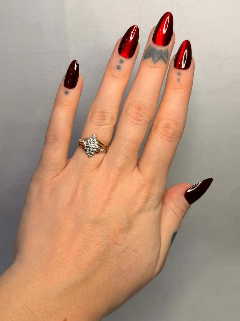 May include: A hand with long, oval-shaped, glossy red nails. A gold ring with a diamond cluster is on the ring finger. The nails have a deep red color, and the ring has a gold band.
