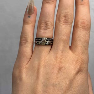 May include: Two rings are displayed, one with a diamond and the other with a black band and a decorative pattern. The diamond ring has a silver band with a small diamond. The other ring has a black band with a repeating pattern.