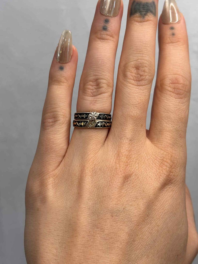 May include: Two rings are displayed, one with a diamond and the other with a black band and a decorative pattern. The diamond ring has a silver band with a small diamond. The other ring has a black band with a repeating pattern.