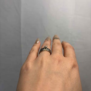 May include: A silver ring with a small, clear gemstone at its centre. The band has a black, patterned design. The ring is on a finger, highlighting the intricate details of the jewellery.