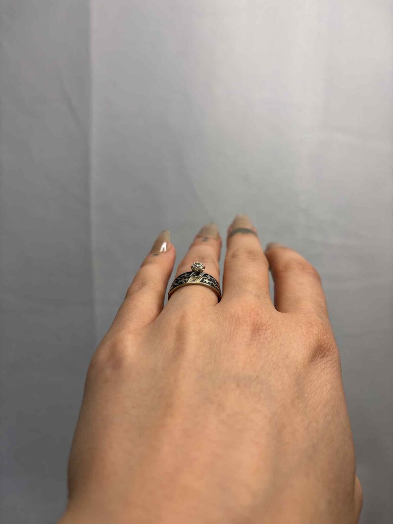 May include: A silver ring with a small, clear gemstone at its centre. The band has a black, patterned design. The ring is on a finger, highlighting the intricate details of the jewellery.
