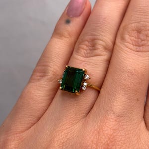 May include: A gold ring featuring a large, emerald-cut, deep green gemstone. The ring is accented with small, round, clear gemstones. The ring is set on a gold band.