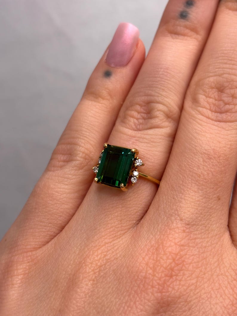 May include: A gold ring featuring a large, emerald-cut, deep green gemstone. The ring is accented with small, round, clear gemstones. The ring is set on a gold band.