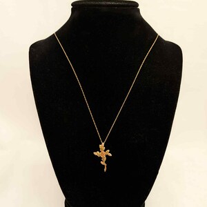 Pode incluir: Um colar dourado com um pingente de cruz floral &eacute; exibido em um manequim preto. O pingente apresenta um design de rosa e folhas. O colar &eacute; suspenso por uma corrente delicada.