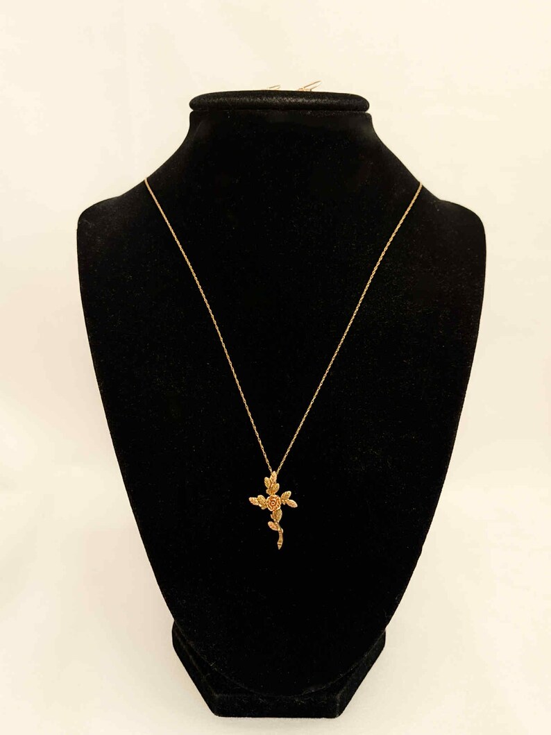 Pode incluir: Um colar dourado com um pingente de cruz floral &eacute; exibido em um manequim preto. O pingente apresenta um design de rosa e folhas. O colar &eacute; suspenso por uma corrente delicada.