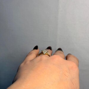 May include: A gold engagement ring with a cluster of small diamonds forming a heart shape. The ring is on a hand with dark nail polish. The band has smaller diamonds. The background is a plain gray.