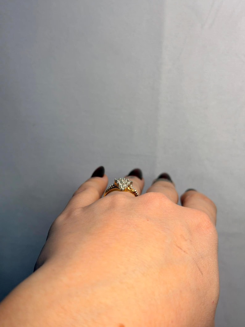 May include: A gold engagement ring with a cluster of small diamonds forming a heart shape. The ring is on a hand with dark nail polish. The band has smaller diamonds. The background is a plain gray.
