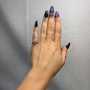 May include: A hand with long, iridescent, coffin-shaped nails in shades of green, blue, and purple. A small, ornate ring with red gemstones adorns the ring finger. Small tattoos are visible on the fingers.