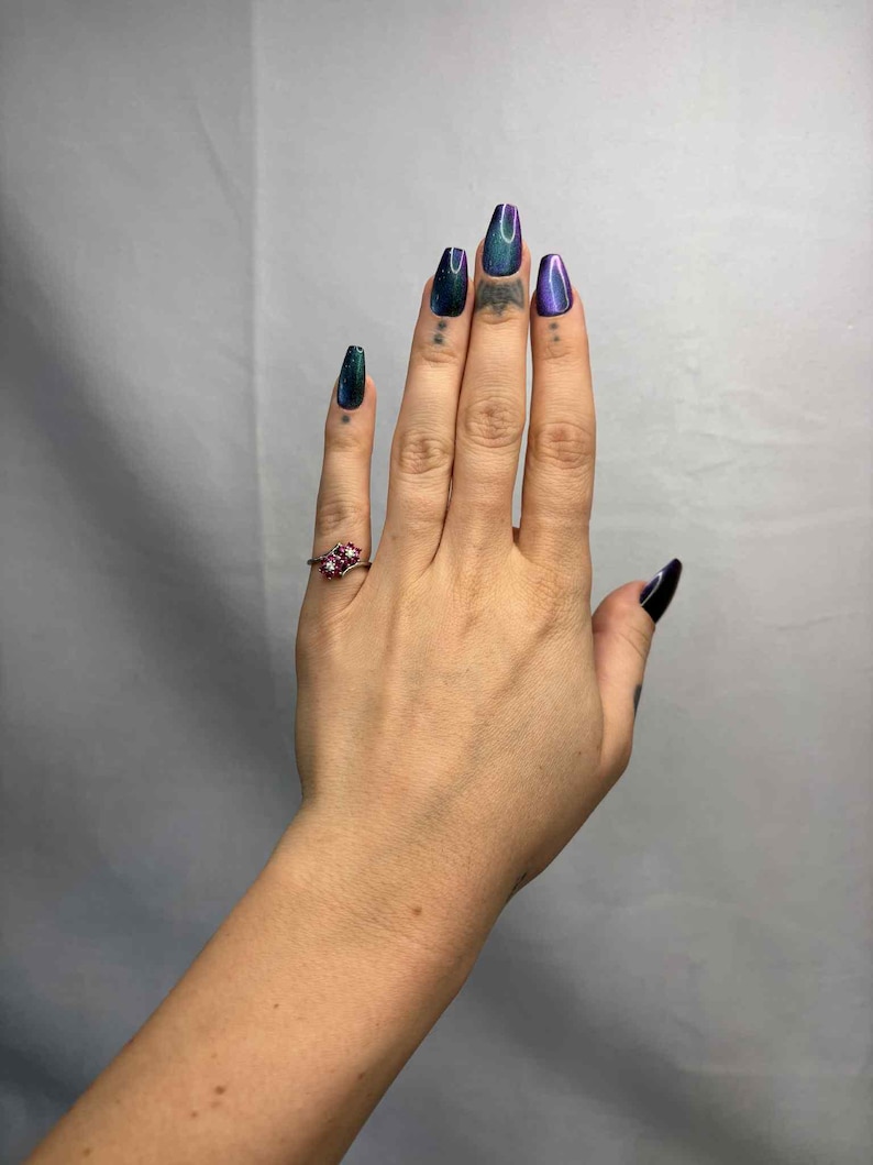 May include: A hand with long, iridescent, coffin-shaped nails in shades of green, blue, and purple. A small, ornate ring with red gemstones adorns the ring finger. Small tattoos are visible on the fingers.