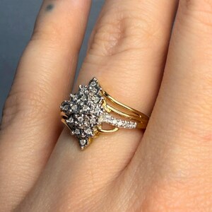 May include: A gold ring with a cluster of small, clear gemstones. The ring has a split shank design with additional small gemstones. The ring is worn on a finger with a dark red nail polish.
