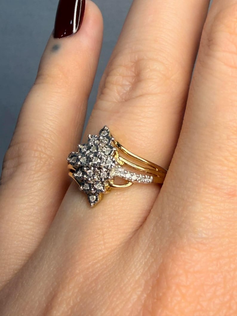 May include: A gold ring with a cluster of small, clear gemstones. The ring has a split shank design with additional small gemstones. The ring is worn on a finger with a dark red nail polish.