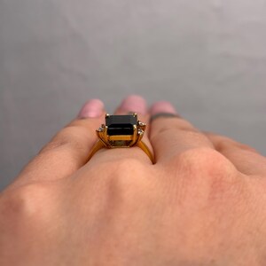 May include: A gold ring featuring a large, rectangular black gemstone as the centerpiece. The ring has a classic design with smaller accent stones on either side of the main stone. The band is a polished gold color.