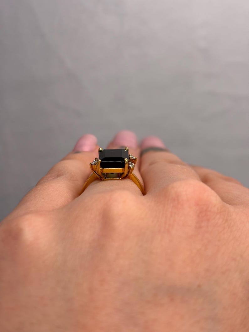 May include: A gold ring featuring a large, rectangular black gemstone as the centerpiece. The ring has a classic design with smaller accent stones on either side of the main stone. The band is a polished gold color.
